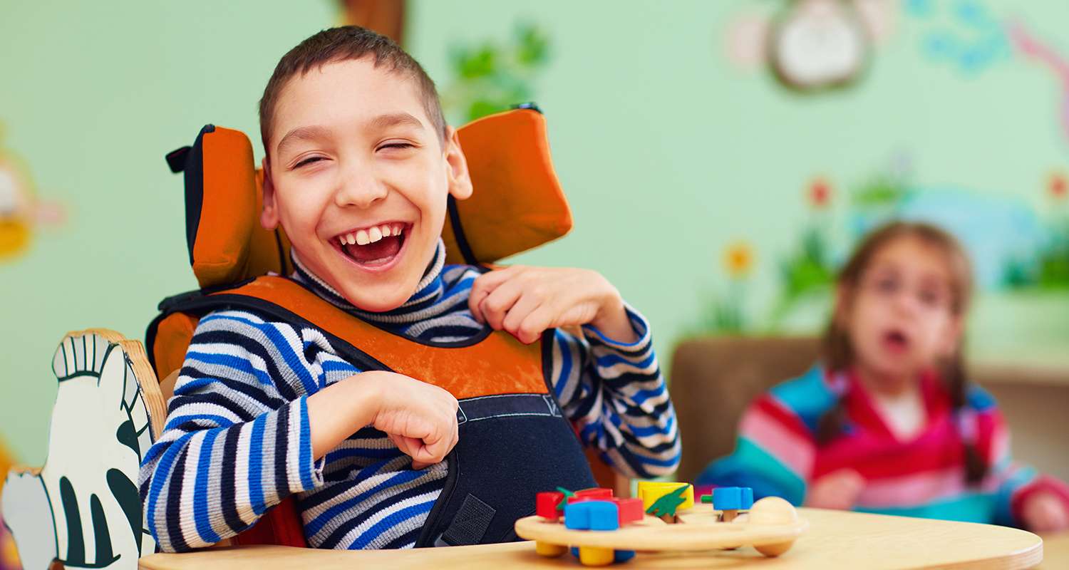 Happy-young-special-needs-child-in-his-harness-chair-at-a-table-in-a-classroom