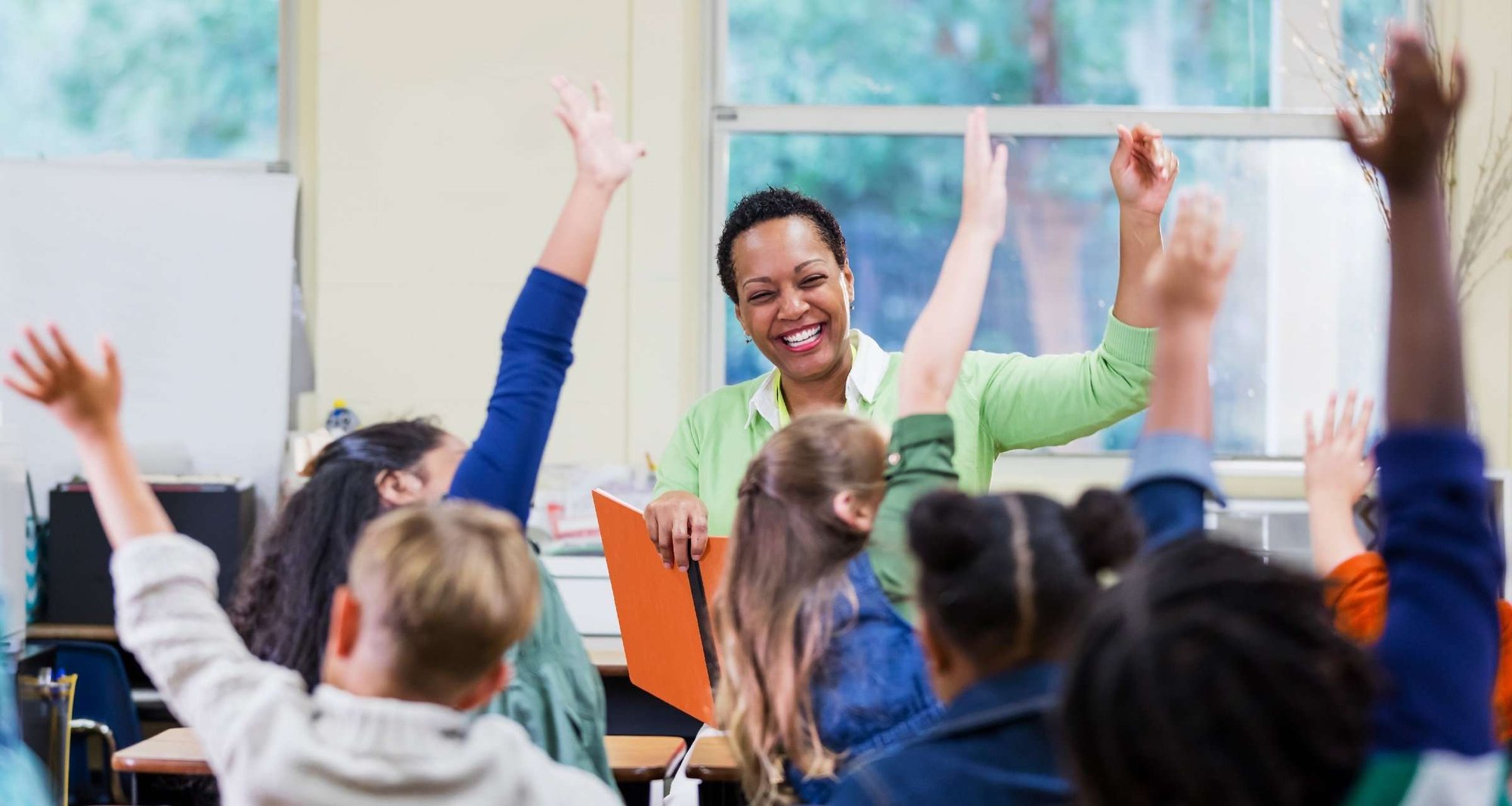 Teacher in front of classroom of elementary students raising their hands.