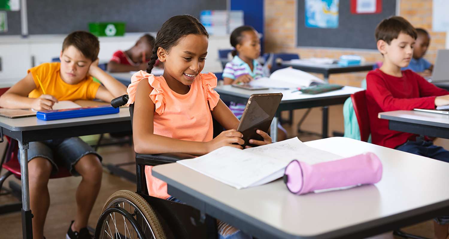 Young-girl-in-a-wheelchair-at-her-desk-reading-on-a-tablet-in-class