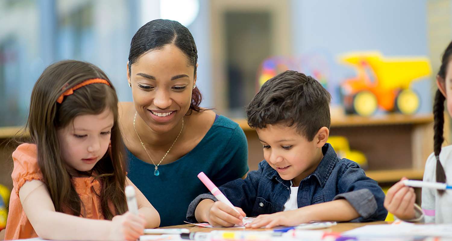 Teacher-sitting-at-table-with-2-young-children-watching-them-color-with-markers