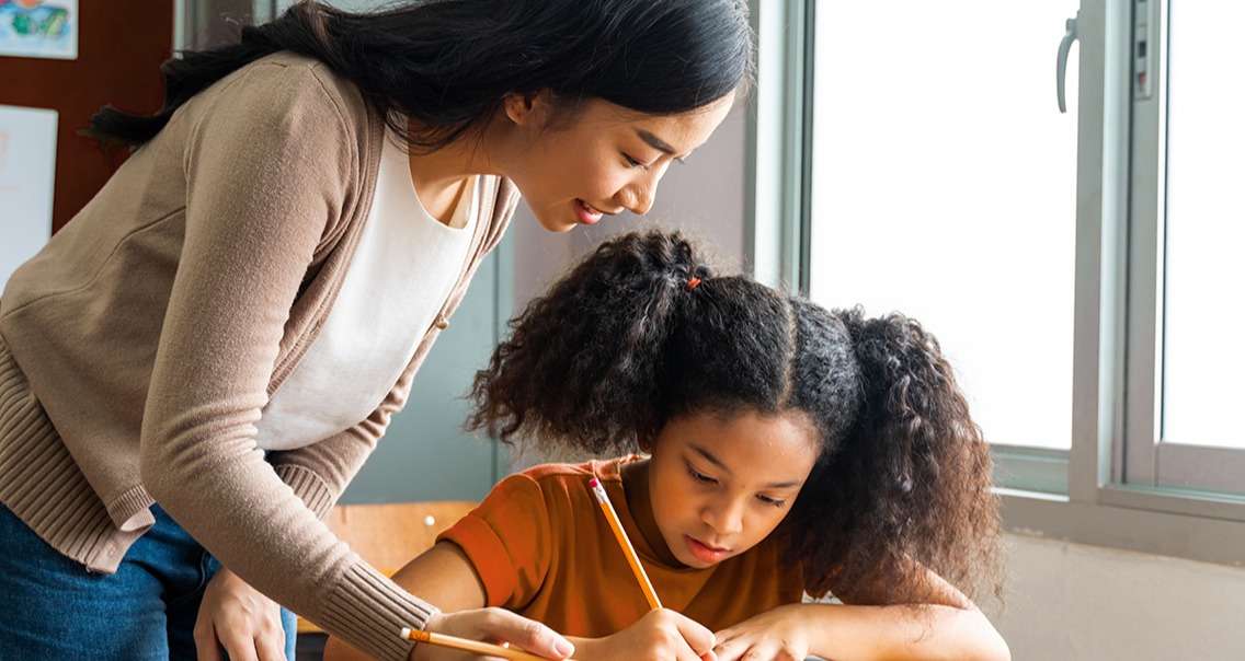 Elementary-age-student-writing-on-paper-at-a-desk-with-help-from-teacher